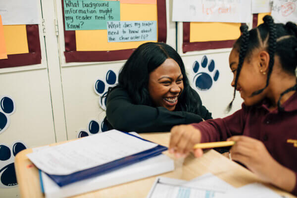 Teacher kneeling at a desk, laughing with a scholar — reference photo from achievementfirst.org