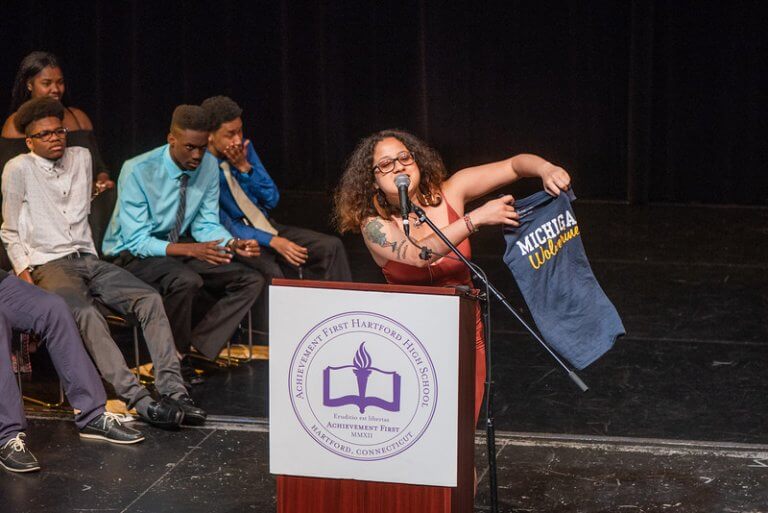 Thalía speaking at a podium holding up a t-shirt from the University of Michigan